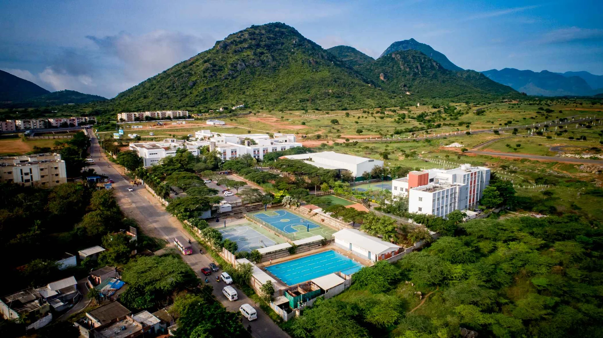 Aerial view of Academy Coimbatore School campus featuring sports courts, swimming pool, and buildings against lush green mountains