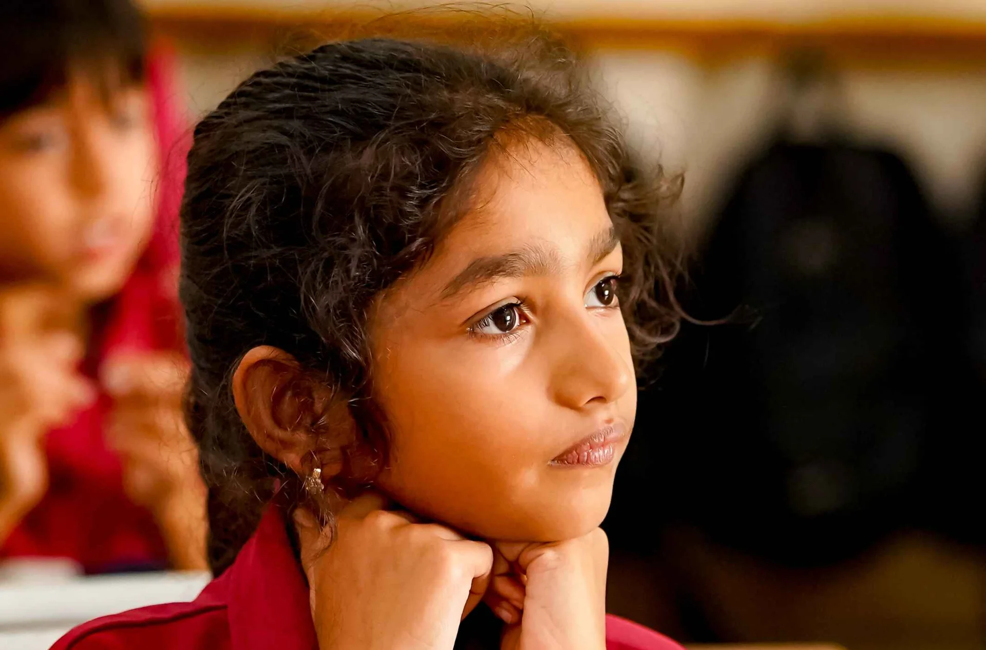 Young student in maroon school uniform resting chin on hands, attentively listening in classroom with workbook on desk