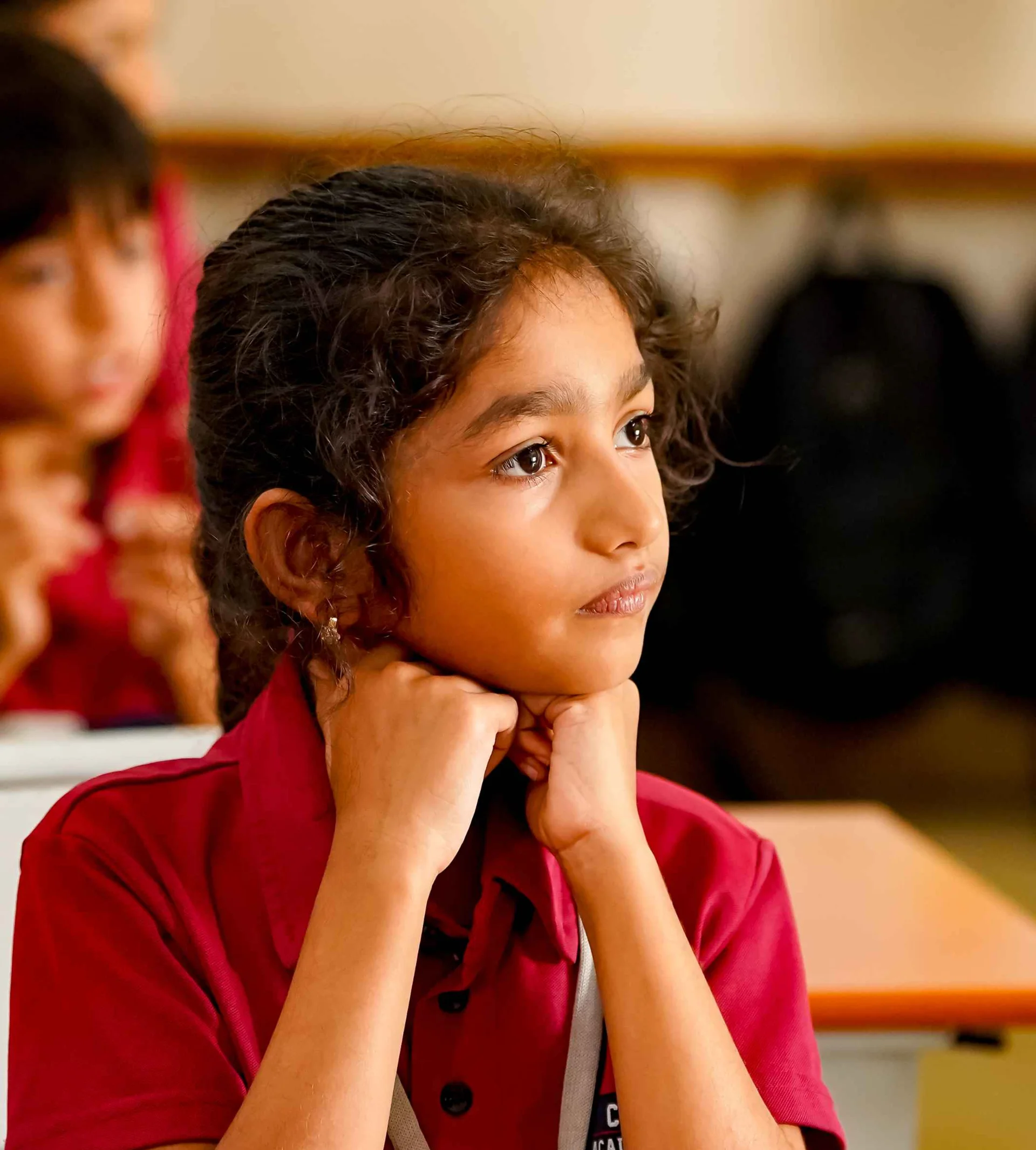 Young student in maroon school uniform resting chin on hands, attentively listening in classroom with workbook on desk