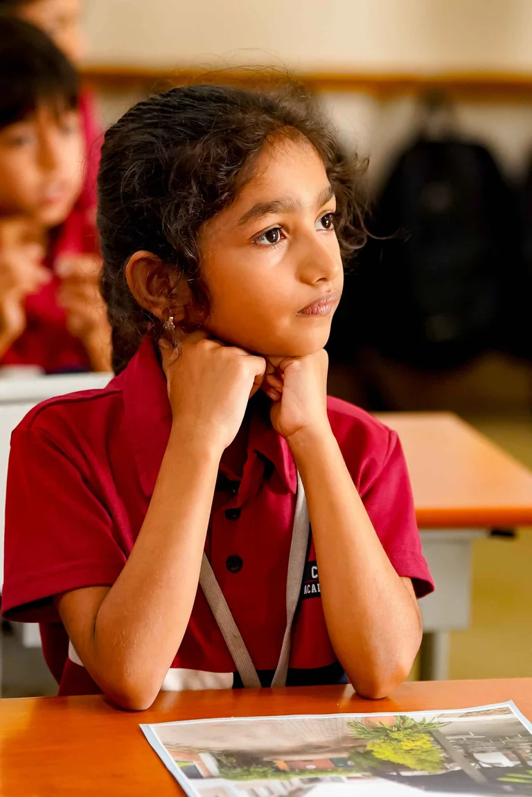 Young student in maroon school uniform resting chin on hands, attentively listening in classroom with workbook on desk