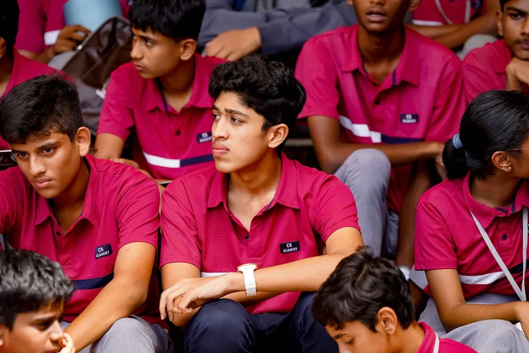 Students in magenta polo shirts with CS Academy logo seated in rows, attentively watching an event or assembly