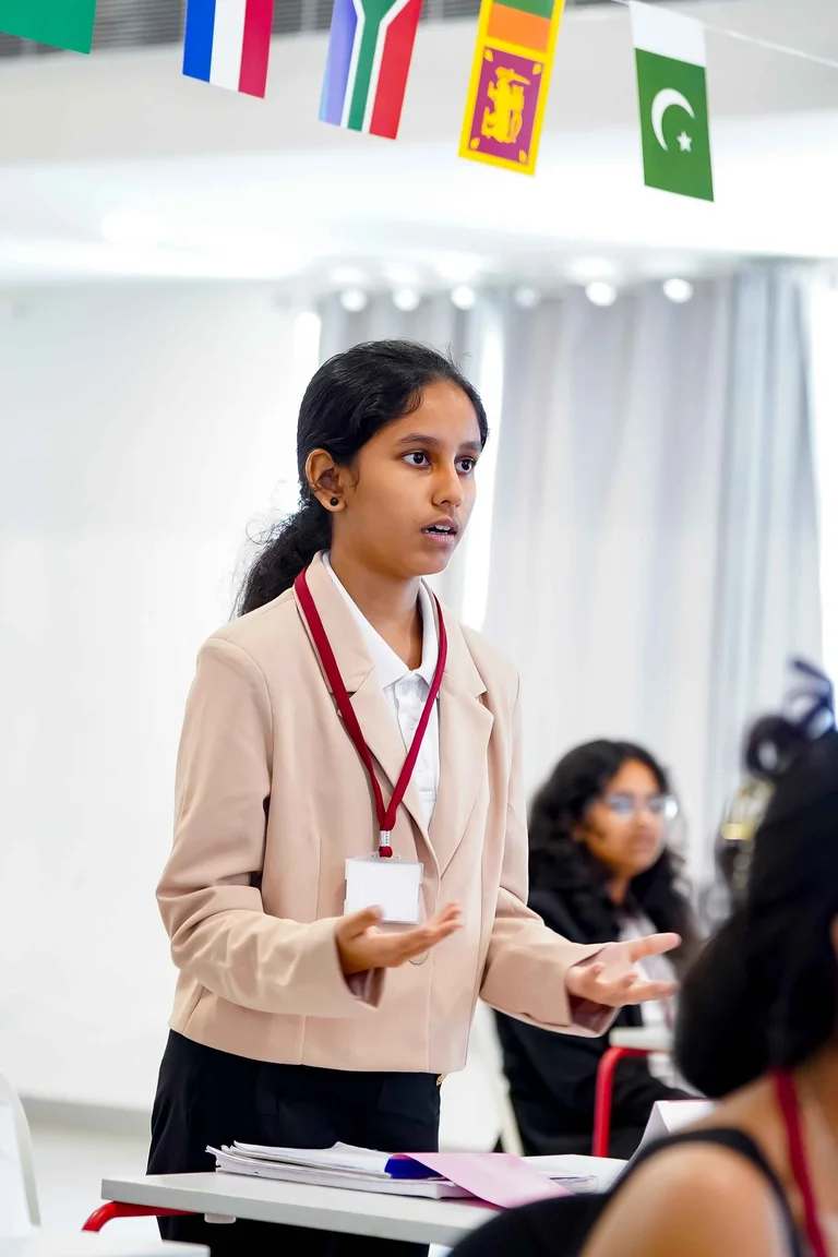 Student in blazer speaking and gesturing during Model United Nations debate with international flags hanging overhead