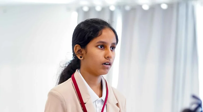 Student in blazer speaking and gesturing during Model United Nations debate with international flags hanging overhead