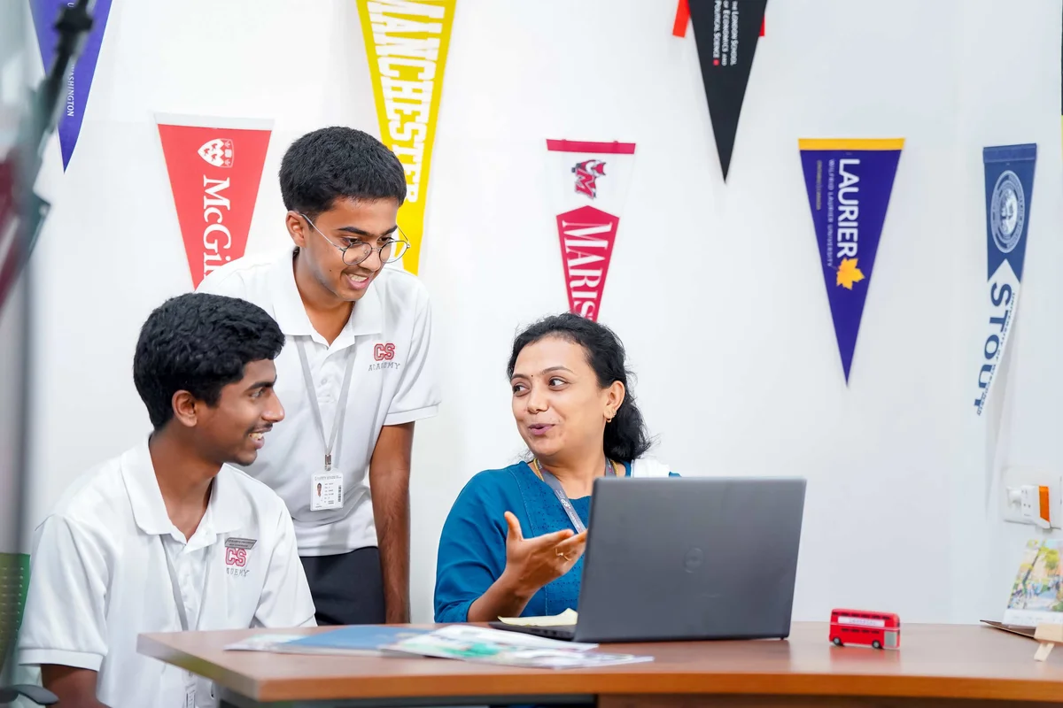 Counselor explaining university options to two students on laptop, with college pennants including McGill and Laurier on wall