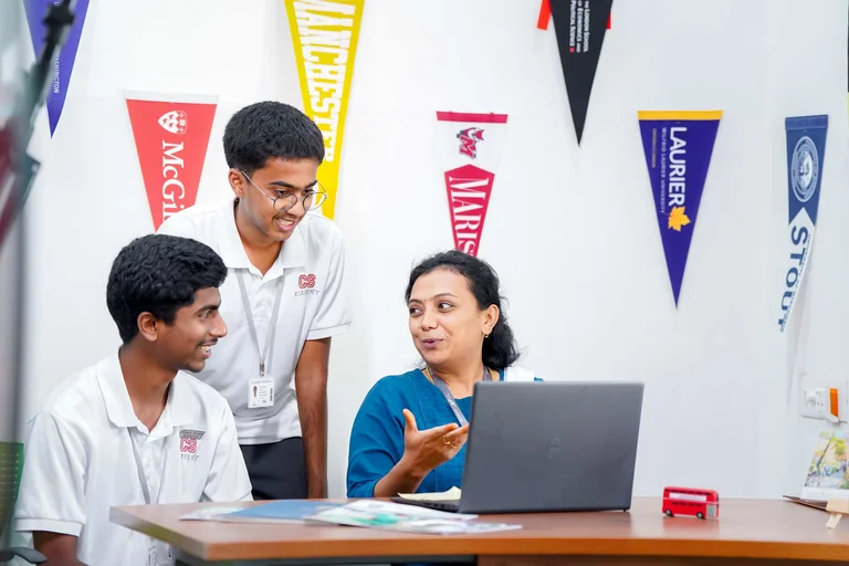 Counselor explaining university options to two students on laptop, with college pennants including McGill and Laurier on wall