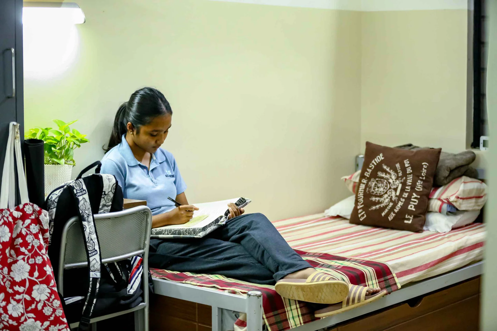 Student in uniform studying with notebook and pen on bed in dormitory room with plant and backpack nearby