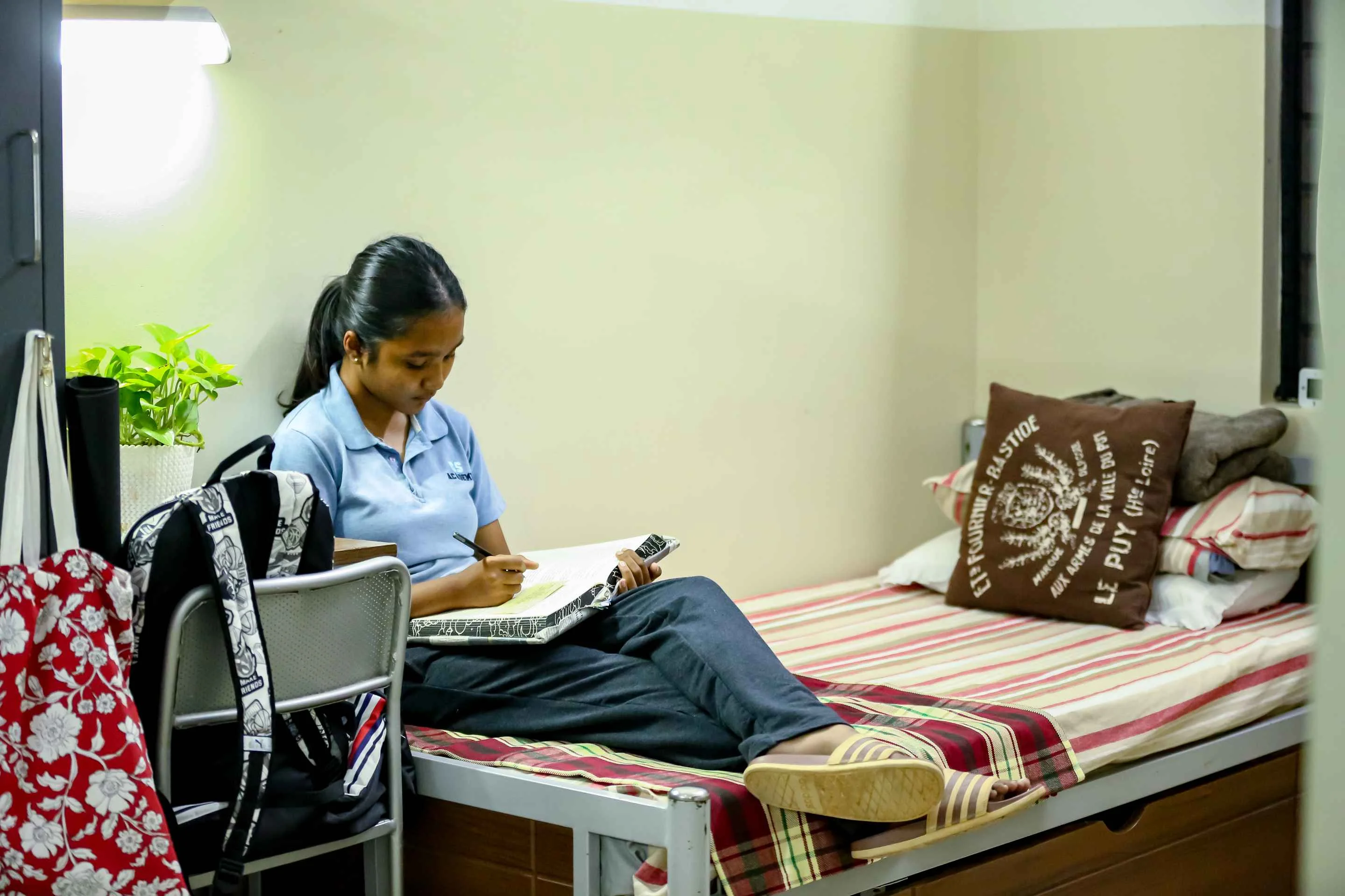 Student in uniform studying with notebook and pen on bed in dormitory room with plant and backpack nearby